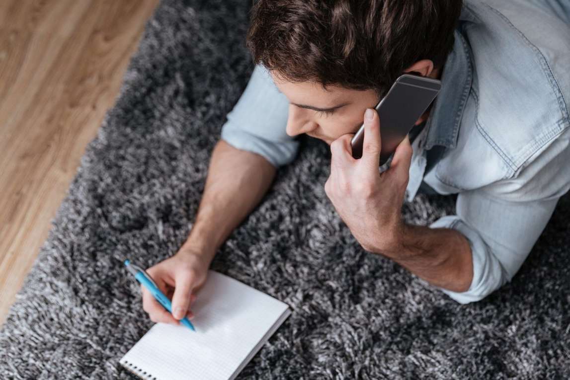 Man writing down questions on the phone laying on a fluffy grey carpet