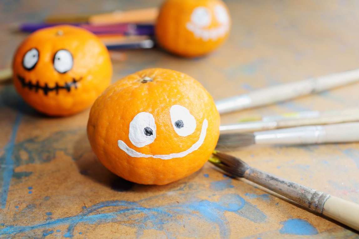 Three pumpkins with hand-painted faces beside paintbrushes for a DIY Halloween decoration project