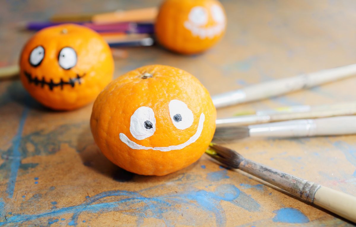 Three pumpkins with hand-painted faces beside paintbrushes for a DIY Halloween decoration project