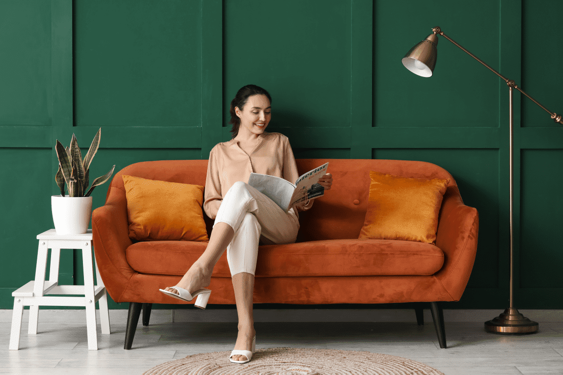 woman reading a magazine sitting on an orange couch with suede upholstery against a green background