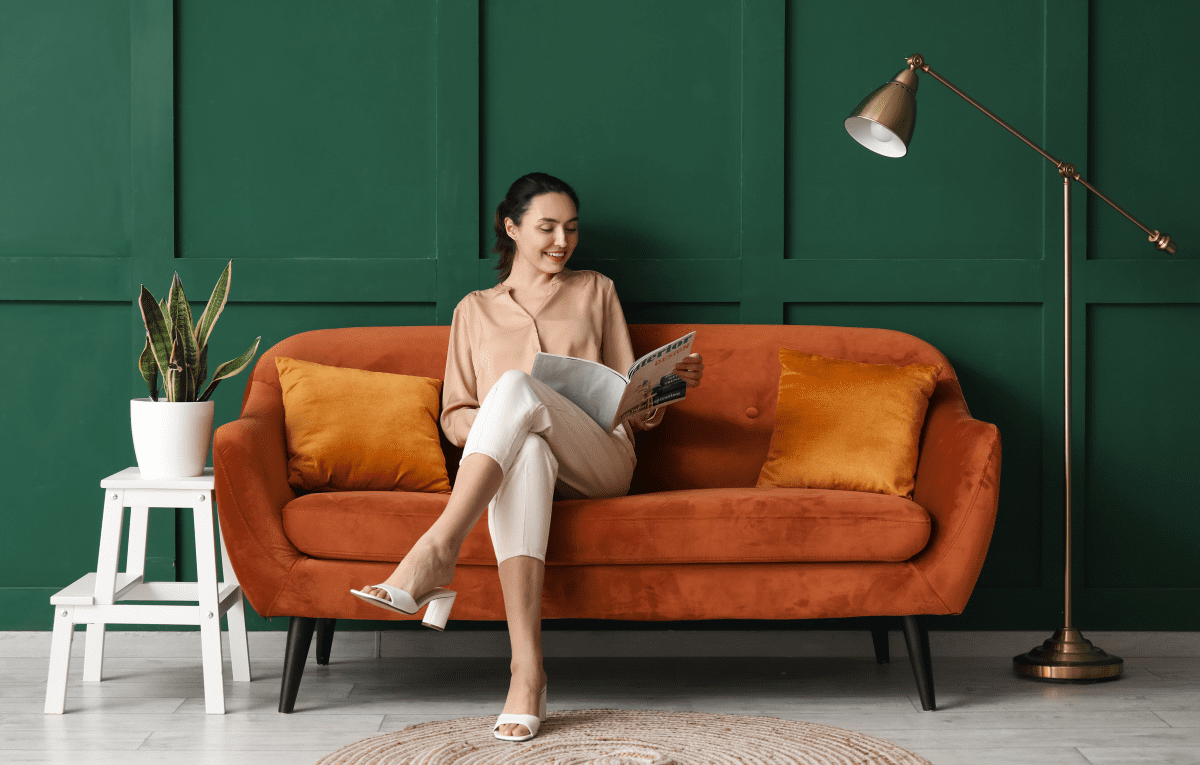 woman reading a magazine sitting on an orange couch with suede upholstery against a green background